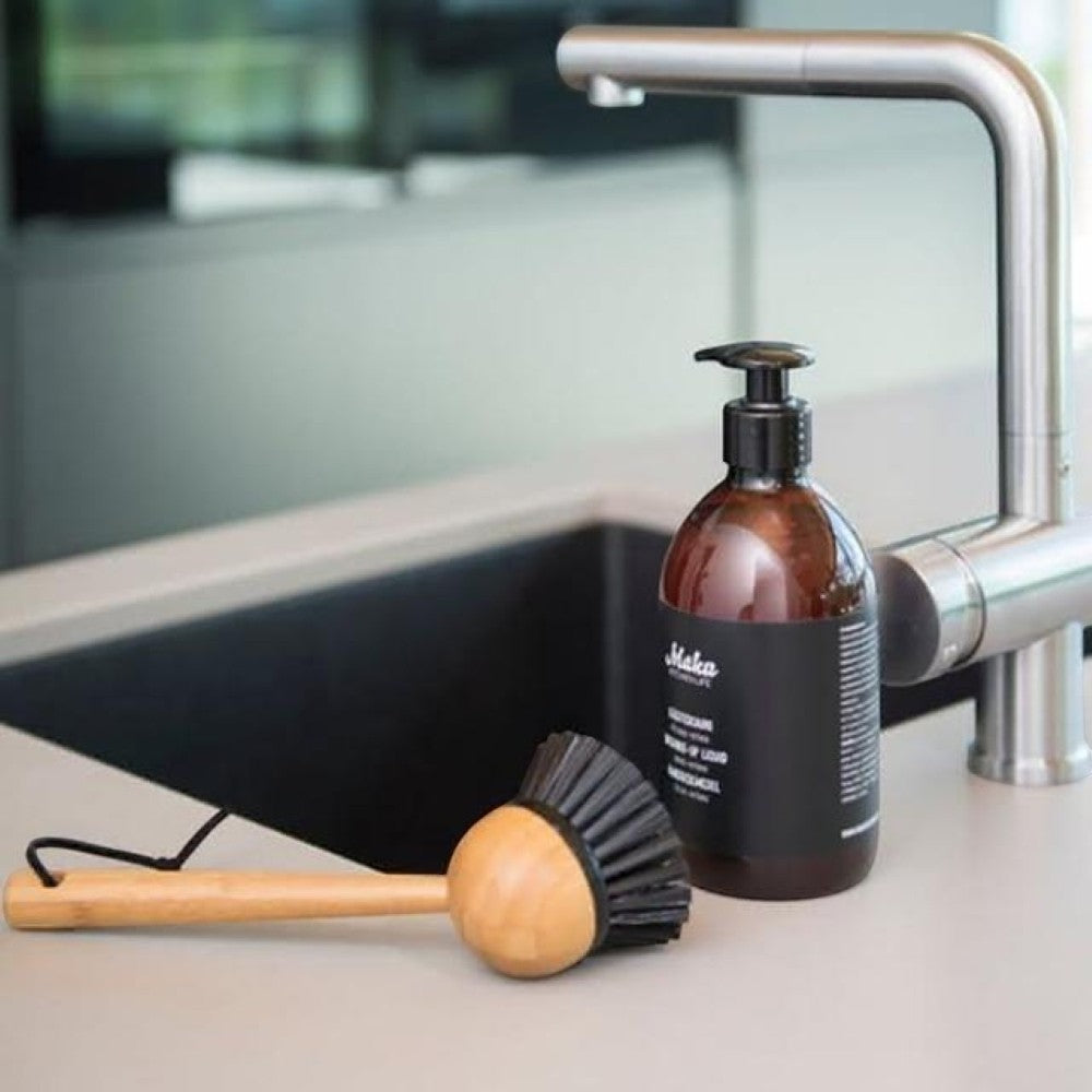 aelgmark bamboo dish brush with black bristles and hanging loop next to a soap dispenser by a kitchen sink.