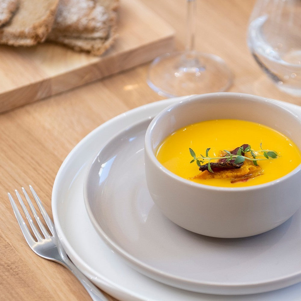 Aelgmark beige ceramic plate with a white bowl of yellow soup topped with herbs and crispy bits.