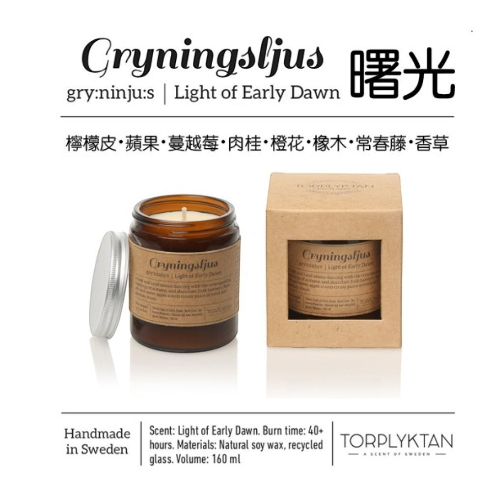 aelgmark amber glass candle with silver lid, featuring a 'Light of Early Dawn' scent with notes of citrus, oak, and apple.