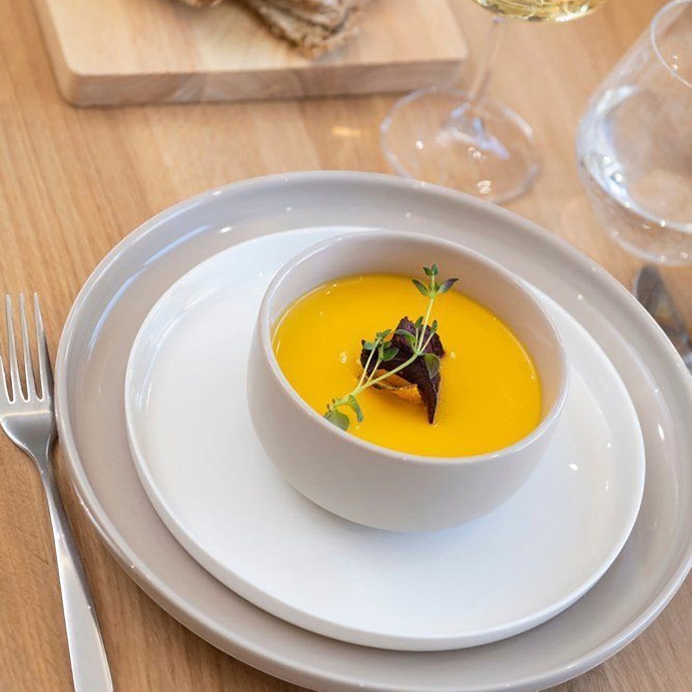 Aelgmark beige ceramic dinner plate and white plate with a beige soup bowl filled with yellow soup topped with herbs.