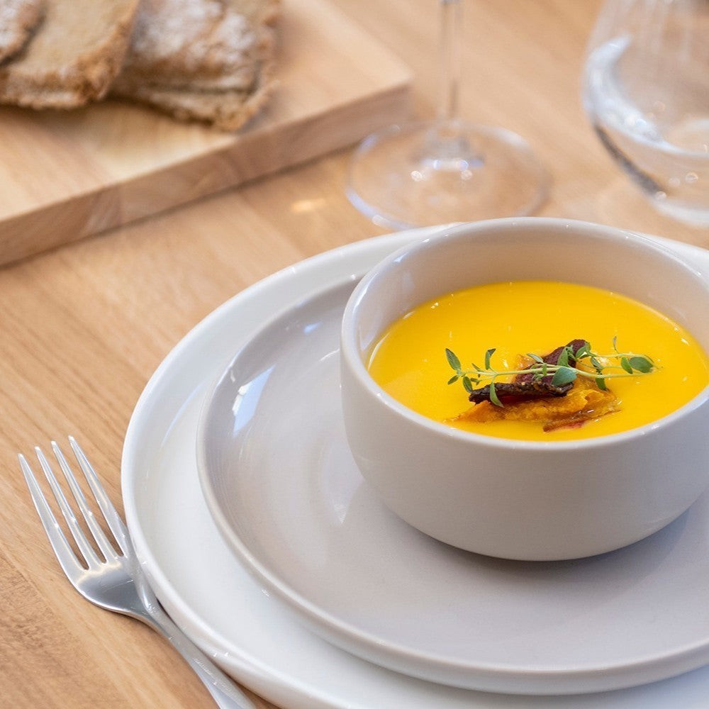 Aelgmark beige ceramic plate with a white bowl of yellow soup topped with herbs and crispy bits.