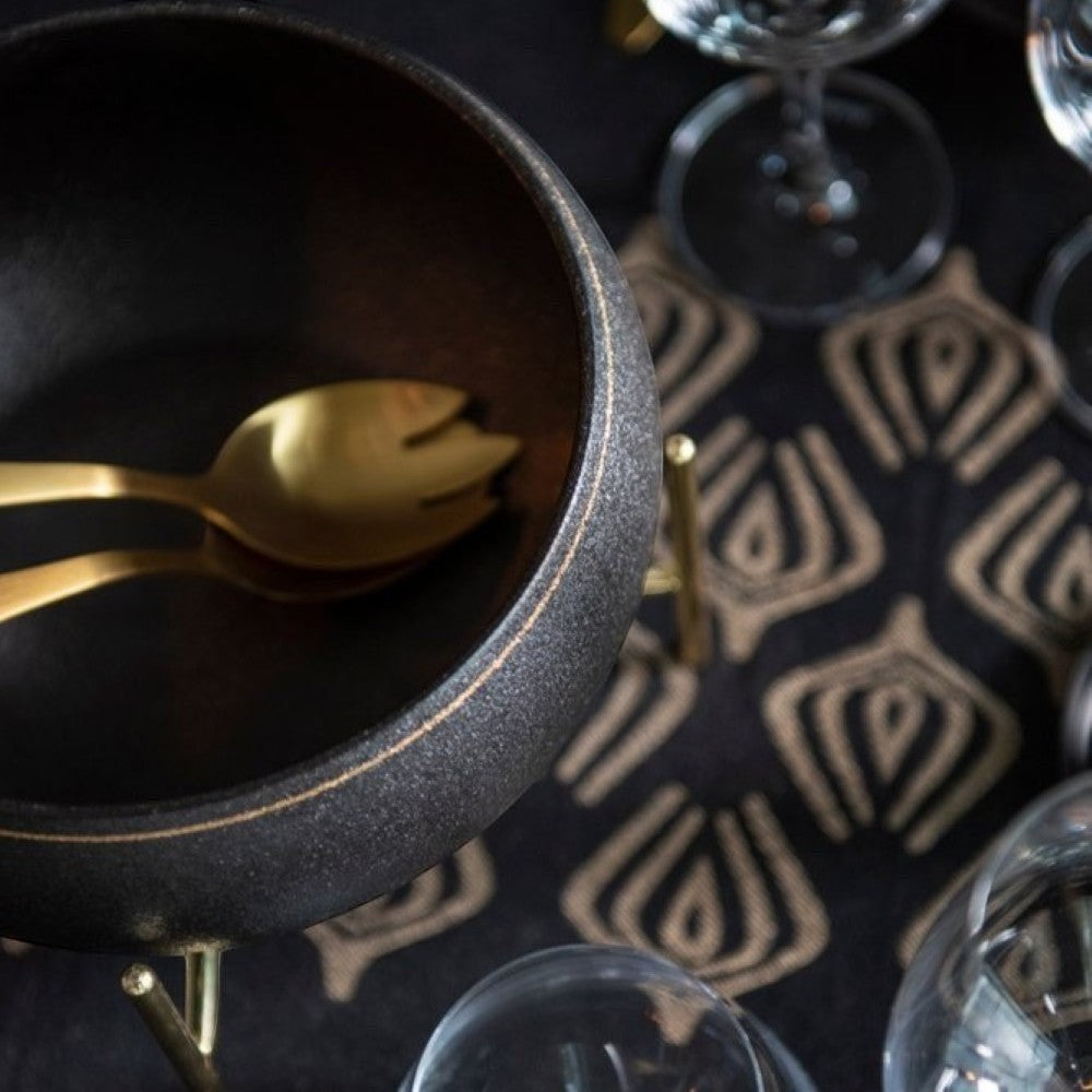 aelgmark black textured salad bowl with gold trim and gold salad servers inside, set on a patterned runner with wine glasses.