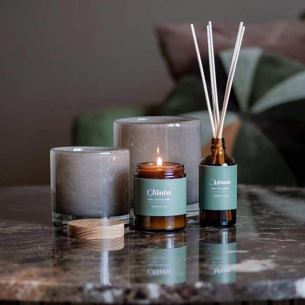 aelgmark Glade scented candle in amber glass jar with green label, beside a glass diffuser and decorative vase.