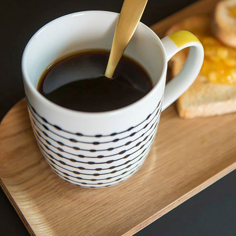 Aelgmark gold spoon in a white mug with black line pattern and yellow handle, serving coffee and toast.