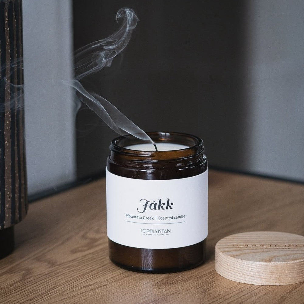 aelgmark Mountain Creek scented candle in amber glass jar with white label, smoke rising from wick.