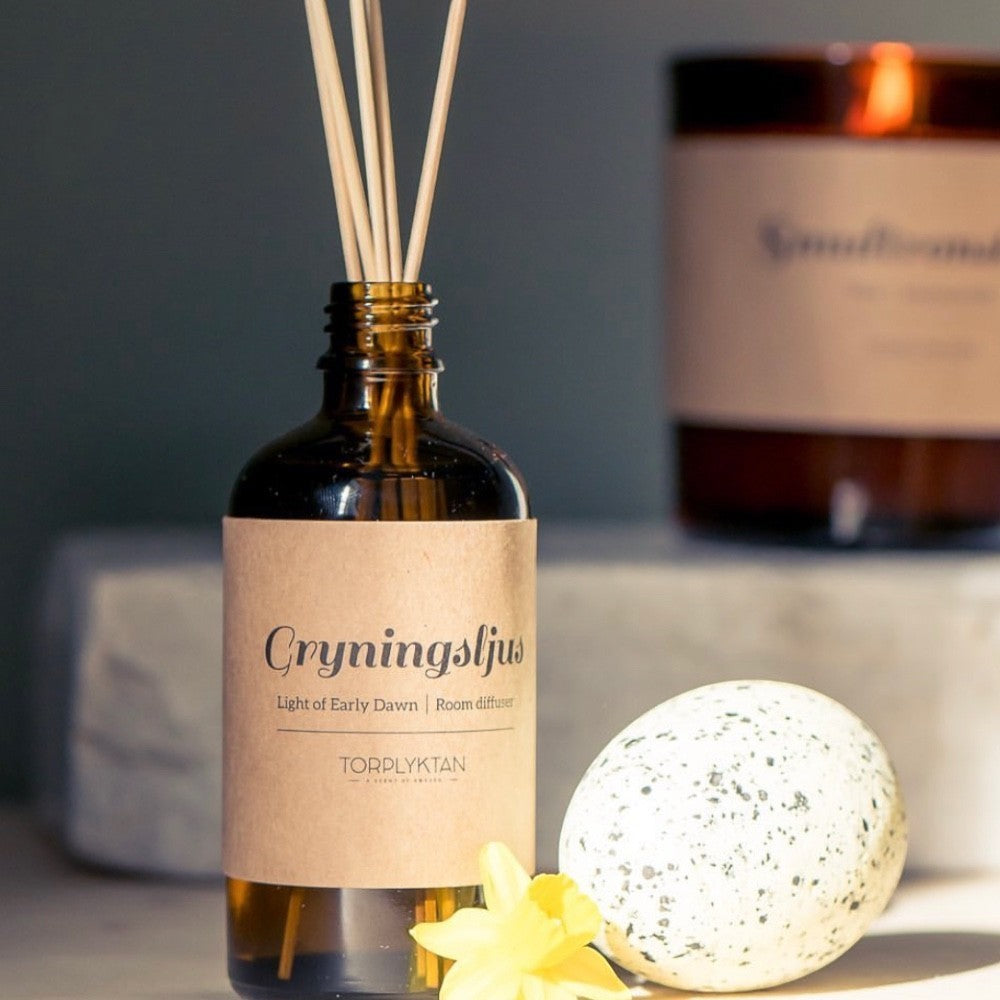 aelgmark amber glass room diffuser with reeds, featuring 'Light of Early Dawn' scent, with a decorative speckled egg and yellow flower.