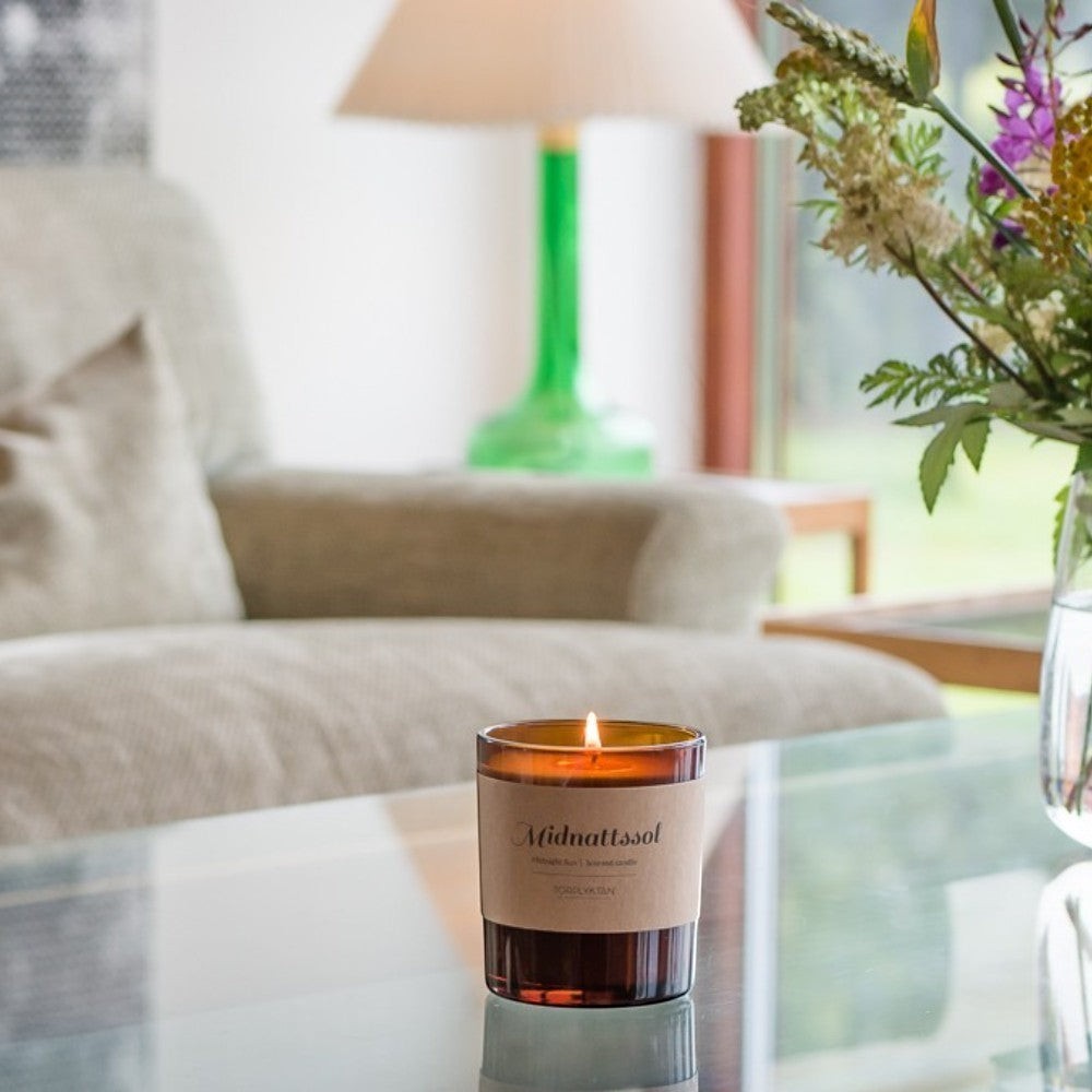 aelgmark Scandinavian scented candle in amber glass with a kraft paper label, lit flame, on a glass table with a sofa and flowers in the background.