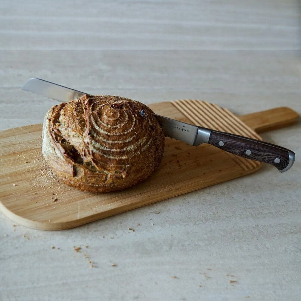 aelgmark 北歐生活選物 oval bamboo bread board with groove detail and a serrated knife cutting a loaf of sourdough bread.
