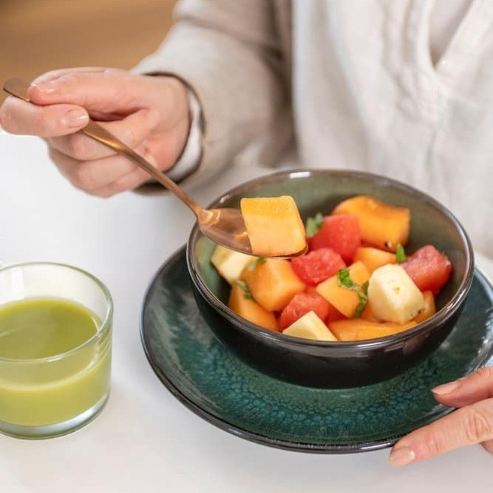 Aelgmark rose gold stainless steel spoon lifts a piece of cantaloupe from a fruit salad in a dark bowl.