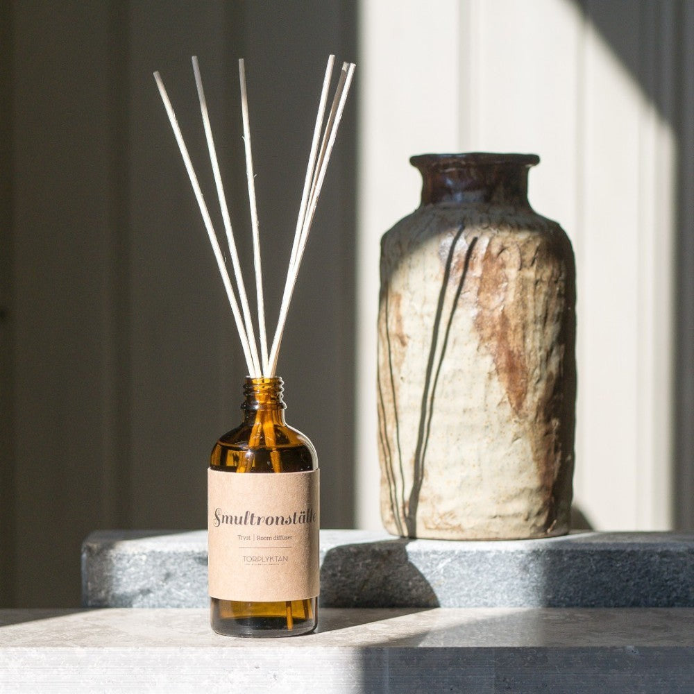 aelgmark amber glass reed diffuser with white reeds, featuring a brown paper label with text. Next to it is a rustic ceramic vase.