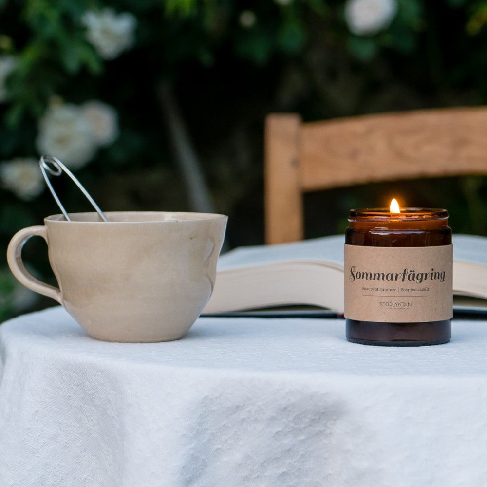 aelgmark北歐生活選物: Torplyktan 'Beauty of Summer' scented candle in amber glass jar with lit flame, beside a mug and open book.