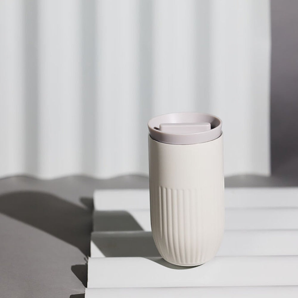 aelgmark Tova travel mug in off-white ceramic with ribbed texture and a light grey lid. Double-wall vacuum insulated.