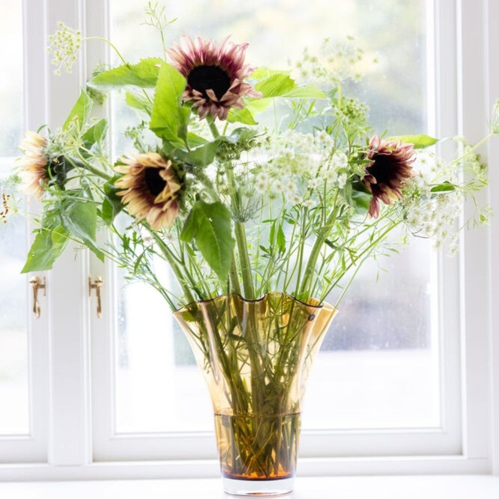 aelgmark Viva amber glass vase with ruffled rim, holding sunflowers and greenery. Nordic design.